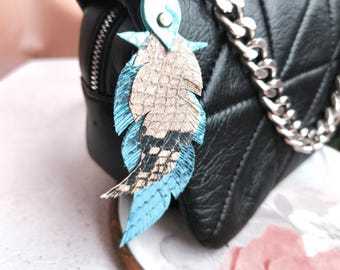 May include: Black quilted leather handbag with silver chain strap. A blue and tan leather feather charm hangs from the chain. The charm is attached with a silver ring. The background is blurred.