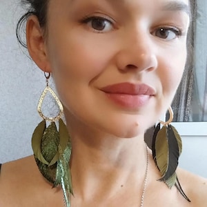 May include: A pair of long, dangling earrings with green and gold feathers. The earrings feature a gold teardrop-shaped pendant with a cut-out design.