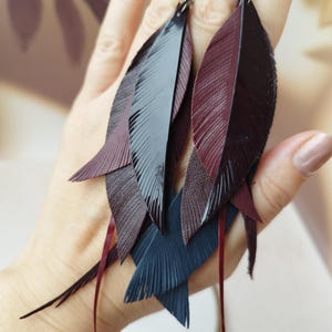 May include: Dangle earrings featuring layered feather-shaped cutouts in shades of deep burgundy, glossy black, and dark navy blue. The earrings are constructed from thin, flexible material with fine etched lines mimicking natural feather textures.