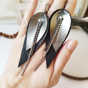 Puede incluir: Un par de pendientes colgantes con un diseño en capas. Presentan una base de cuero negro, una capa metálica plateada y una cadena colgante de pequeños y brillantes pedrería. Los pendientes están diseñados con un cierre de gancho.