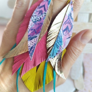 Puede incluir: Un par de aretes largos de plumas coloridas con detalles dorados. Los aretes están hechos de cuero en capas y flecos en tonos de rosa, amarillo, turquesa y azul. Las plumas están adornadas con un patrón metálico dorado.