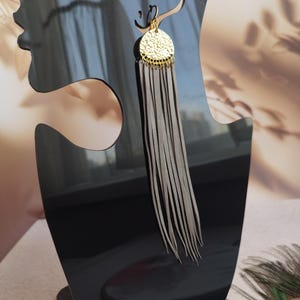 May include: A single gold-colored earring with a circular, textured pendant and long, gray leather fringe. The earring is displayed on a black mannequin head, showcasing its design and length. The earring's design is suitable for a variety of occasions.