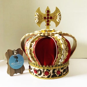 May include: A decorative crown with a red velvet interior, gold accents, and a leopard print base. Adorned with seashells and red jewels, it features a gold emblem on top. The crown is likely for ceremonial or decorative purposes.