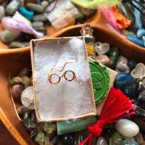 May include: A gold-colored ring in the shape of Harry Potter's glasses, displayed in a small white box with a brown lid. The box is surrounded by colorful stones and beads.