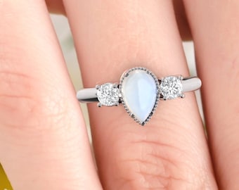 May include: A pearlescent teardrop-shaped gemstone ring with two smaller round gemstones on either side. The ring is set in silver.