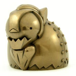 May include: A metallic bronze-coloured sculpture of a stylised creature with a smooth, rounded body, a jagged jawline, and a row of small, raised circular studs along its curved back. The object features a solid, uniform finish throughout.