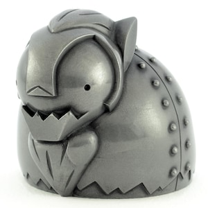 May include: A metallic grey, rounded figurine featuring a stylised creature with a pointed beard, jagged mouth, and small circular eyes. The back section of the object displays a curved panel adorned with a vertical row of raised, rivet-like studs.