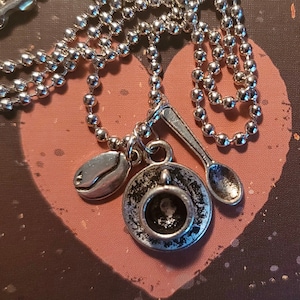 May include: A silver necklace with a charm of a coffee cup and saucer, a coffee bean, and a spoon. The necklace is on a brown background with a heart shape.