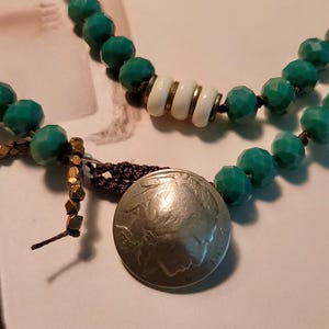 May include: A green and white beaded necklace with a silver coin pendant. The coin has a profile of a Native American on it.
