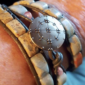 May include: A multi-strand bracelet with a silver charm featuring a circular design with a cross and dots. The bracelet is made of brown leather and light brown, blue, and pink stones.