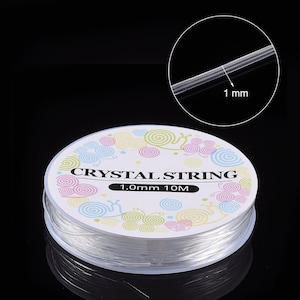 Peut inclure: Une bobine de fil de cristal transparent, de 1,0 mm d'&eacute;paisseur et de 10 m&egrave;tres de long, est pr&eacute;sent&eacute;e sur fond noir. L'&eacute;tiquette porte l'inscription "CRYSTAL STRING" et des motifs en spirale color&eacute;s.