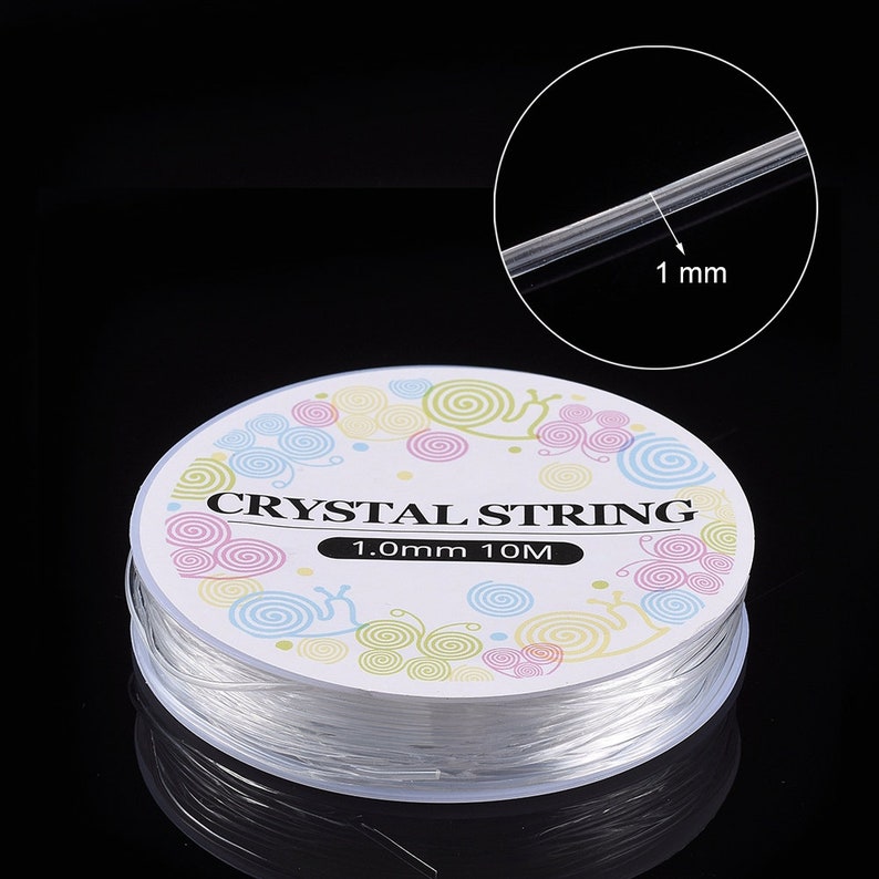 Peut inclure: Une bobine de fil de cristal transparent, de 1,0 mm d'&eacute;paisseur et de 10 m&egrave;tres de long, est pr&eacute;sent&eacute;e sur fond noir. L'&eacute;tiquette porte l'inscription "CRYSTAL STRING" et des motifs en spirale color&eacute;s.