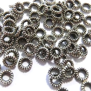 Può includere: Un mucchio di anelli di salto in argento, ognuno con un design testurizzato e a spirale. Gli anelli sono sparsi su uno sfondo bianco.