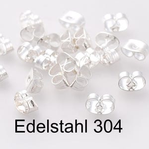 Könnte beinhalten: Nahaufnahme einer Ansammlung von silbernen Ohrringverschlüssen. Die Verschlüsse sind aus Edelstahl und haben einen Schmetterlingsverschluss. Der Text "Edelstahl 304" ist auf dem Bild sichtbar.