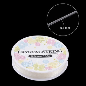 Peut inclure: Une bobine de fil de cristal transparent, de 0,6 mm d'&eacute;paisseur et de 15 m&egrave;tres de long, est pr&eacute;sent&eacute;e sur fond noir. L'&eacute;tiquette indique "CRYSTAL STRING" avec un motif tourbillonnant d&eacute;coratif.