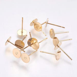 Könnte beinhalten: Eine Sammlung goldfarbener Ohrringstecker mit flachen, runden Pads. Die Stifte sind dünn und gerade, mit einer kleinen, kreisförmigen Scheibe am Ende zum Befestigen von Ohrringen. Die Pads haben eine leicht erhöhte Mitte.