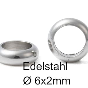 Può includere: Due anelli in metallo argentato, ciascuno con un bordo arrotondato e una finitura liscia e lucida. Gli anelli hanno un diametro di 6 mm e uno spessore di 2 mm. Il testo "Edelstahl Ø 6x2mm" è stampato sotto gli anelli.