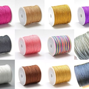 Könnte beinhalten: Eine Auswahl an farbigen Kordelspulen in verschiedenen Farbtönen wie Pink, Braun, Gold, Lila, Schwarz, Koralle und Weiß. Die Kordeln sind eng auf weißen Kunststoffspulen aufgewickelt, ideal für Bastelarbeiten und Schmuckherstellung.