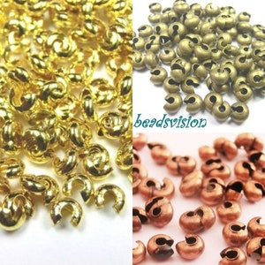 May include: A close-up of three piles of gold, bronze, and copper crimp beads. The beads are small and round with a hole in the centre. They are used for jewellery making.