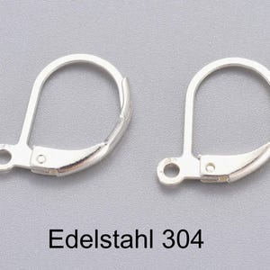 Könnte beinhalten: Zwei silberfarbene Ohrringverschlüsse. Jeder Verschluss hat eine gebogene Oberseite und einen Klapphebel mit einer kleinen Schlaufe. Der Text "Edelstahl 304" ist darunter gedruckt.