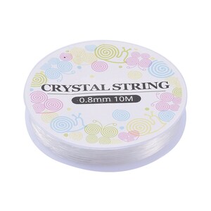 Peut inclure: Bobine de fil de cristal transparent, 0,8 mm d'&eacute;paisseur et 10 m&egrave;tres de long. L'&eacute;tiquette porte l'inscription "CRYSTAL STRING" avec des motifs de spirales et d'escargots aux couleurs pastel. Le fil est bien enroul&eacute;.