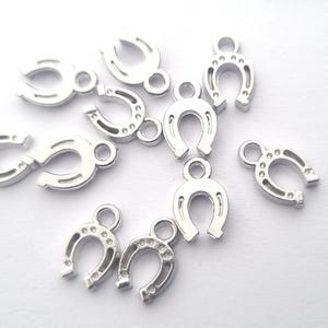 Könnte beinhalten: Eine Sammlung kleiner, silberfarbener Hufeisen-Anhänger. Jeder Anhänger hat oben eine Schlaufe zum Befestigen an Schmuck oder anderen Bastelarbeiten. Die Hufeisen sind mit Rillen und kleinen Löchern versehen, was ihnen ein realistisches Aussehen verleiht.