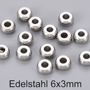 Könnte beinhalten: Nahaufnahme einer Gruppe von 14 silbernen Metallperlen, jede mit einem Loch in der Mitte. Die Perlen haben einen Durchmesser von 6 mm und eine Höhe von 3 mm. Der Text "Edelstahl 6x3mm" ist unter den Perlen sichtbar.