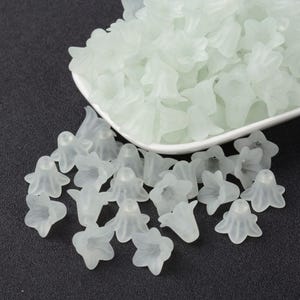 Könnte beinhalten: Eine Schale gefüllt mit weißen, blumenförmigen Perlen. Die Perlen sind durchscheinend und haben eine matte Oberfläche. Die Perlen sind auf einer schwarzen Oberfläche verstreut.