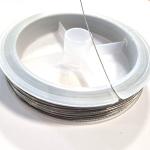 Op de afbeelding: Een spoel dun, zilverkleurig draad. Het draad is strak gewikkeld rond een witte plastic spoel met een centrale naaf en een heldere, cirkelvormige buitenrand. Het draad loopt van de spoel af, wat suggereert dat het klaar is voor gebruik.