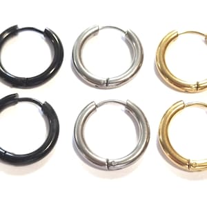 Könnte beinhalten: Sechs Creolen mit Scharnier, in einer Reihe angeordnet, in Schwarz, Silber und Gold. Die Ohrringe haben ein dickes, kreisförmiges Design mit glatter, polierter Oberfläche. Die Ohrringe werden vor einem weißen Hintergrund präsentiert. Jeder Ohrring hat einen Durchmesser von ca. 2 cm.
