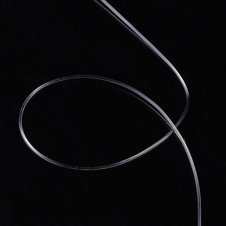 Peut inclure: Un filament de plastique clair, fin et flexible, possiblement un c&acirc;ble &agrave; fibre optique, se courbe et se croise sur un fond noir. Le mat&eacute;riau semble lisse et r&eacute;fl&eacute;chissant, avec des reflets subtils.