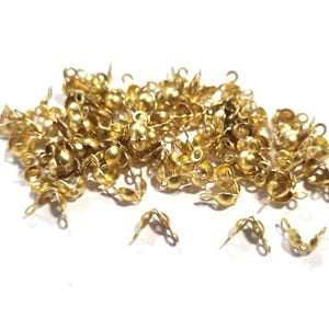 Könnte beinhalten: Eine Ansammlung kleiner, goldfarbener Perlenkappen und -verbinder, verstreut auf einer weißen Oberfläche. Die Perlenkappen haben ein blütenblattartiges Design, und die Verbinder haben eine Schlaufe zur Befestigung. Ideal für die Schmuckherstellung.