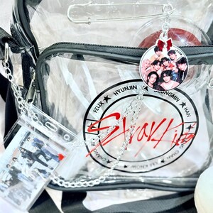 Puede incluir: Una bolsa de pl&aacute;stico transparente con una correa negra y una cadena plateada. La bolsa tiene un logotipo en blanco y negro con el texto "Stray Kids" y los nombres de los miembros. Hay un llavero de pl&aacute;stico transparente con una foto de los miembros de la banda unido a la bolsa.