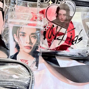 Puede incluir: Una bolsa de pl&aacute;stico transparente con una correa negra y una cadena plateada. La bolsa tiene un dije en forma de coraz&oacute;n rojo con una foto de una persona dentro. La bolsa tambi&eacute;n tiene un bolsillo de pl&aacute;stico transparente con una foto de una persona dentro. La bolsa tiene las palabras "Stray Kids" escritas en ella.