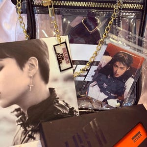 K&ouml;nnte beinhalten: Eine transparente Plastiktasche mit einem goldenen Kettenriemen. Die Tasche enth&auml;lt eine Photocard eines K-Pop-Bandmitglieds, einen kleinen Anh&auml;nger mit dem Text "DELUCO FOR ATEEZ" und eine schwarz-orangefarbene Box mit dem Text "ATEEZ".