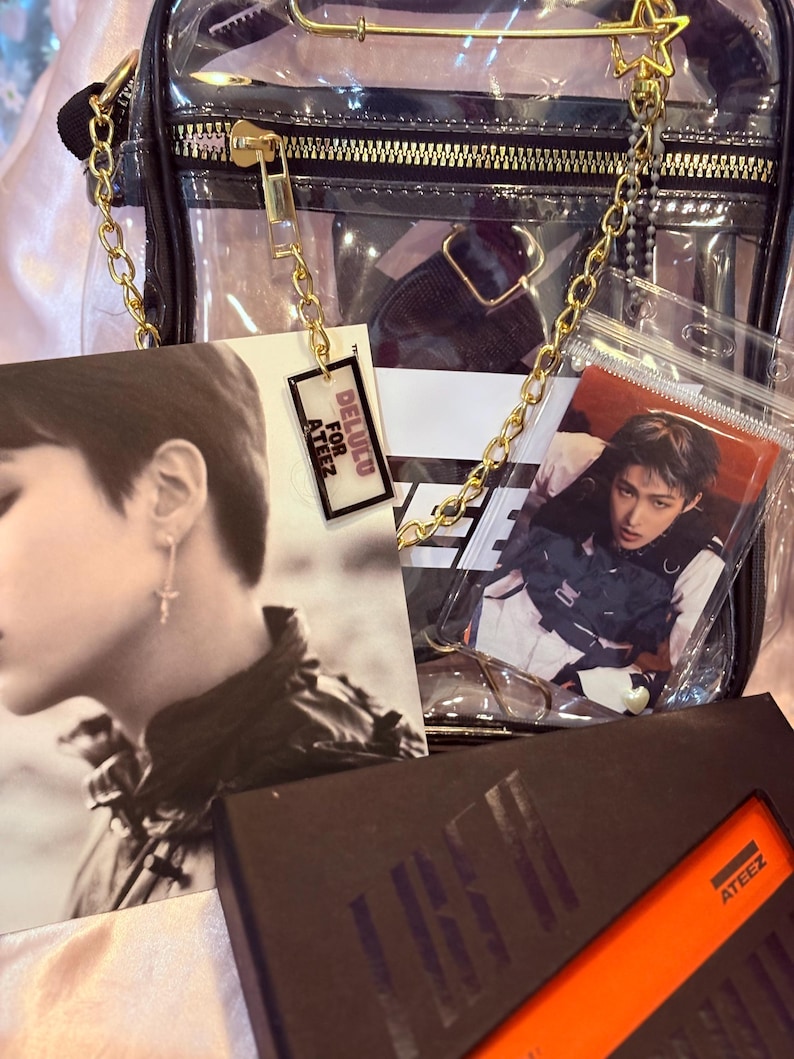 K&ouml;nnte beinhalten: Eine transparente Plastiktasche mit einem goldenen Kettenriemen. Die Tasche enth&auml;lt eine Photocard eines K-Pop-Bandmitglieds, einen kleinen Anh&auml;nger mit dem Text "DELUCO FOR ATEEZ" und eine schwarz-orangefarbene Box mit dem Text "ATEEZ".