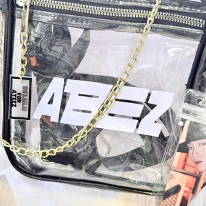 K&ouml;nnte beinhalten: Transparente Plastikt&uuml;te mit einem goldenen Kettenriemen und einem schwarzen Rei&szlig;verschluss. Die Tasche hat ein wei&szlig;es Logo mit der Aufschrift "Ateez" und ein kleines Etikett mit der Aufschrift "Delulu for Ateez".