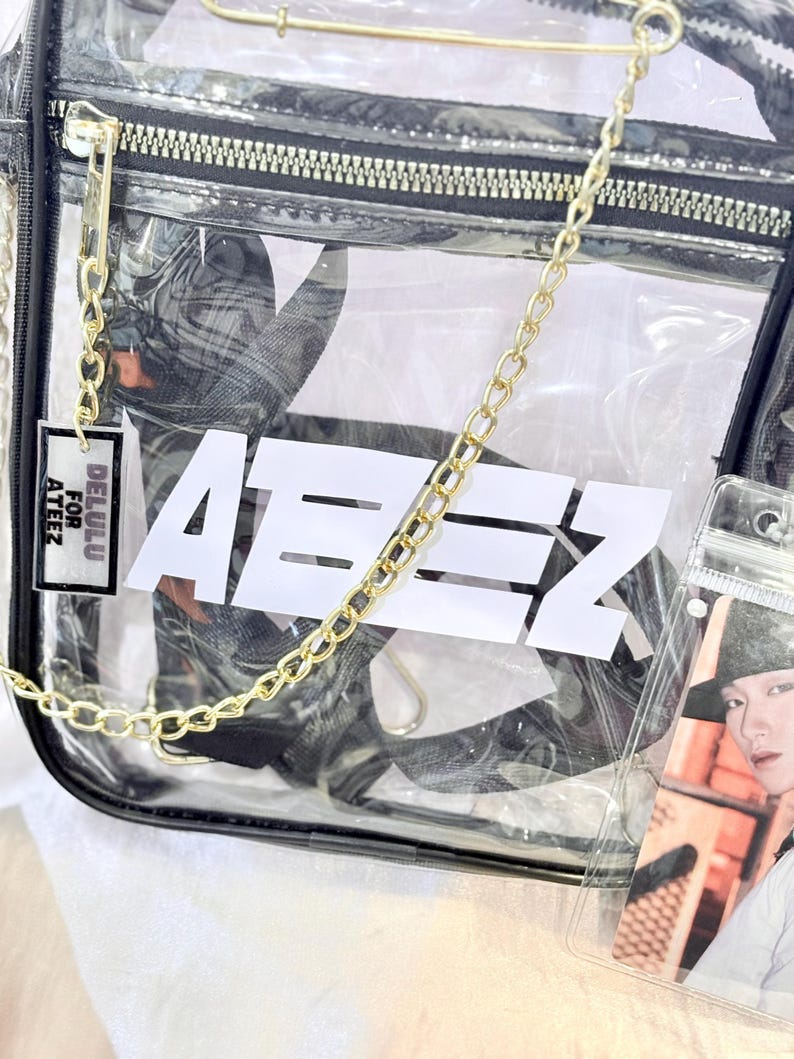 K&ouml;nnte beinhalten: Transparente Plastikt&uuml;te mit einem goldenen Kettenriemen und einem schwarzen Rei&szlig;verschluss. Die Tasche hat ein wei&szlig;es Logo mit der Aufschrift "Ateez" und ein kleines Etikett mit der Aufschrift "Delulu for Ateez".