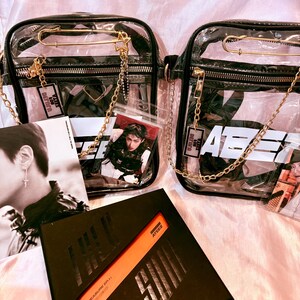 K&ouml;nnte beinhalten: Zwei transparente Plastikt&uuml;ten mit goldenen Ketten und Sicherheitsnadeln. Die Taschen haben schwarze Tr&auml;ger und sind gef&uuml;llt mit Foto-Karten und einem schwarzen Album mit dem Text "TREASURE EP.1: THE FIRST STEP" und "ATEEZ" auf dem Cover.