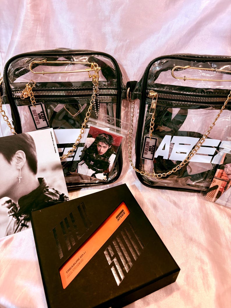 K&ouml;nnte beinhalten: Zwei transparente Plastikt&uuml;ten mit goldenen Ketten und Sicherheitsnadeln. Die Taschen haben schwarze Tr&auml;ger und sind gef&uuml;llt mit Foto-Karten und einem schwarzen Album mit dem Text "TREASURE EP.1: THE FIRST STEP" und "ATEEZ" auf dem Cover.