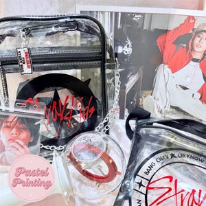 Puede incluir: Bolso transparente de pl&aacute;stico con correa de cadena negra. El bolso tiene una cremallera negra y un logotipo negro y rojo que dice "Stray Kids". El bolso est&aacute; decorado con un peque&ntilde;o amuleto que dice "Hello Stray Kids".