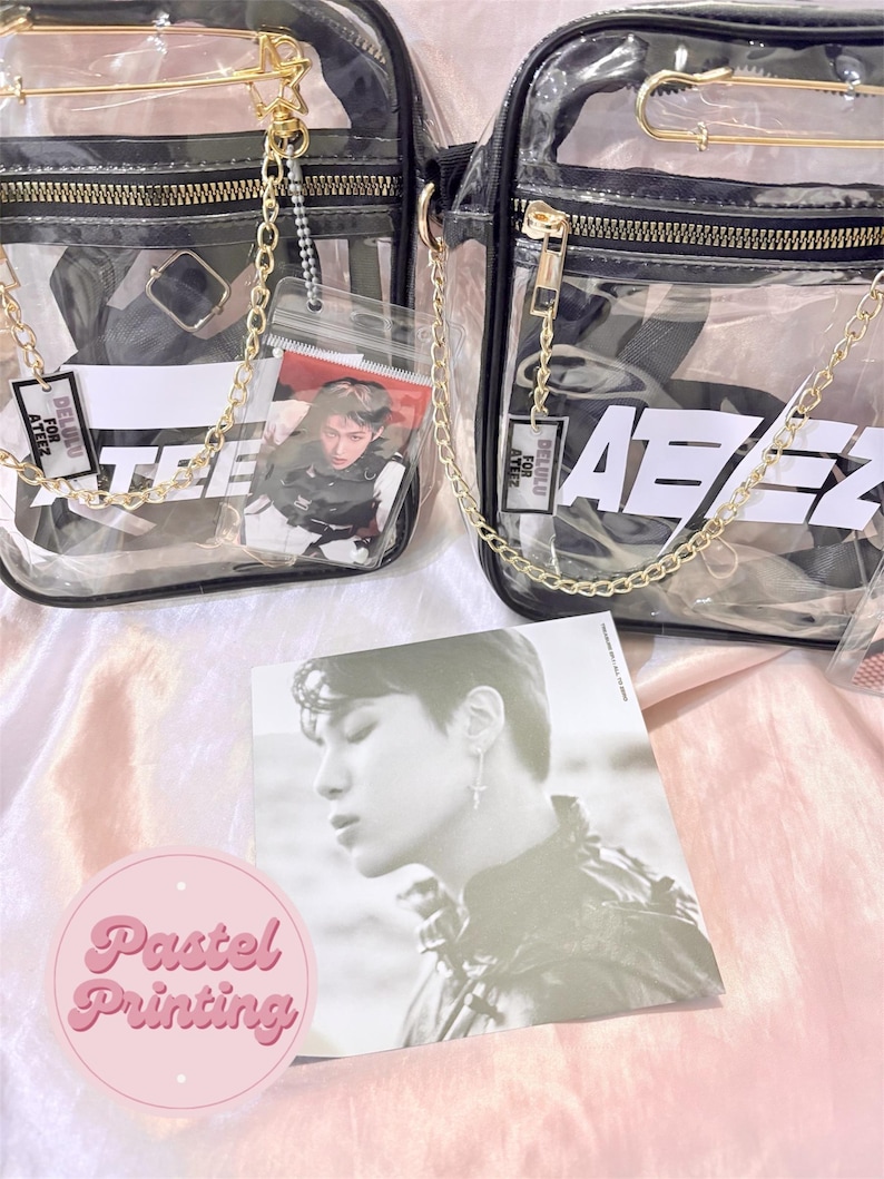 K&ouml;nnte beinhalten: Zwei transparente Plastik-Umh&auml;ngetaschen mit goldenen Ketten und schwarzen Tr&auml;gern. Die Taschen haben wei&szlig;en Text, der "DELUXE FOR ATEEZ" und "ATEEZ" lautet. Die Taschen befinden sich auf einer rosa Oberfl&auml;che mit einer Fotokarte eines Mannes.