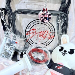 Puede incluir: Una bolsa de pl&aacute;stico transparente con una correa negra y una cadena plateada. La bolsa contiene una varita luminosa blanca, un conejito de peluche blanco con una camiseta negra y roja, y un llavero redondo transparente con una foto de un grupo de K-pop. El llavero est&aacute; unido a la bolsa con una cadena plateada. La bolsa tambi&eacute;n tiene un bolsillo de pl&aacute;stico transparente con una foto del mismo grupo de K-pop. La bolsa tiene un logotipo rojo y blanco que dice "Stray Kids" con los nombres de los miembros alrededor del logotipo.
