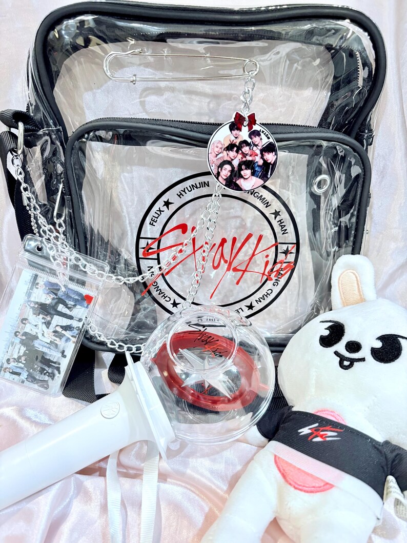 Puede incluir: Una bolsa de pl&aacute;stico transparente con una correa negra y una cadena plateada. La bolsa contiene una varita luminosa blanca, un conejito de peluche blanco con una camiseta negra y roja, y un llavero redondo transparente con una foto de un grupo de K-pop. El llavero est&aacute; unido a la bolsa con una cadena plateada. La bolsa tambi&eacute;n tiene un bolsillo de pl&aacute;stico transparente con una foto del mismo grupo de K-pop. La bolsa tiene un logotipo rojo y blanco que dice "Stray Kids" con los nombres de los miembros alrededor del logotipo.