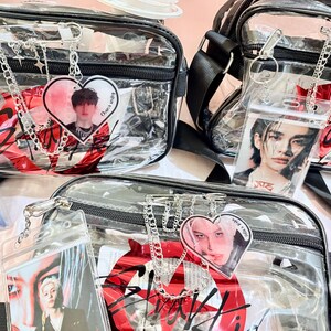 Puede incluir: Tres bolsos de bandolera de pl&aacute;stico transparente con correas y cremalleras negras. Cada bolso tiene un gr&aacute;fico rojo y negro con la palabra "Stray Kids" y un amuleto en forma de coraz&oacute;n con una foto de un miembro diferente del grupo.