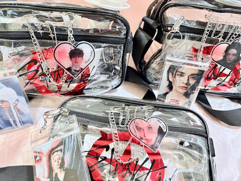 Puede incluir: Tres bolsos de bandolera de pl&aacute;stico transparente con correas y cremalleras negras. Cada bolso tiene un gr&aacute;fico rojo y negro con la palabra "Stray Kids" y un amuleto en forma de coraz&oacute;n con una foto de un miembro diferente del grupo.