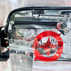 Puede incluir: Bolso transparente de pl&aacute;stico con cierre de cremallera negro y un c&iacute;rculo rojo con el texto "Stray Kids" en negro. El bolso tiene un bolsillo transparente en la parte delantera con una tarjeta de foto en el interior.