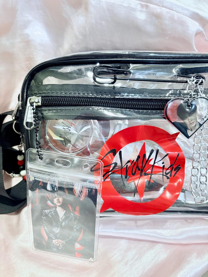 Puede incluir: Bolso transparente de pl&aacute;stico con cierre de cremallera negro y un c&iacute;rculo rojo con el texto "Stray Kids" en negro. El bolso tiene un bolsillo transparente en la parte delantera con una tarjeta de foto en el interior.