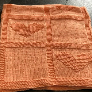 May include: A peach-coloured knitted blanket with a heart design. The blanket is divided into four squares, with two squares featuring a heart shape. The blanket has a textured appearance and a ribbed border.