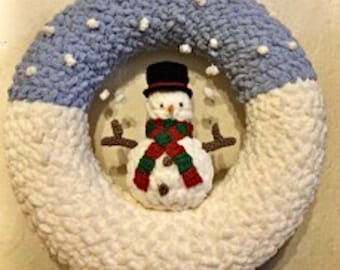 Könnte beinhalten: Ein weißer und blauer gehäkelter Kranz mit einem Schneemann in der Mitte. Der Schneemann trägt einen schwarzen Zylinder, einen grün-roten Schal und hat schwarze Augen und eine Karottennase.